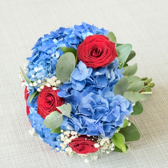 When you lift this bouquet to your chest you can almost feel the sky itself: domed clusters of cobalt-blue hydrangea create a tactile, cloud-like centre, their petal surfaces soft and slightly reflective in natural light. Each deep red rose rises from that blue field with sculptural presence-velvety, full, and saturated with emotion-and the contrast between the two colours gives the arrangement both drama and clarity. Wisps of white gypsophila thread through the composition like embroidered lace, delicate strands that bring an air of innocence and celebration, while sprays of eucalyptus add structural calm with their cool, matte-green leaves and a faint, refreshing scent. The bouquet is constructed to be both handsome and intimate: the hydrangeas provide a plush foundation, roses bring focal intensity, and the smaller accents create visual breathing room so the eye can rest and return. This pairing reads as classic yet contemporary, appropriate for an anniversary, wedding, or any ceremony seeking a refined, patriotic-tinged palette. As prepared by Grove Park Florist, stems are layered with attention to scent and sight-greens tucked to release fragrance without overwhelming the florals-and arranged to withstand handling during a procession or to sit elegantly on a celebratory table. Delivered locally-whether a short walk from Grove Park station or to a lunch at a nearby community garden-the bouquet carries both craft and care. It embodies a feeling of bold love softened by reassurance, a piece meant to be touched, inhaled, and remembered.